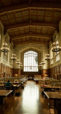 An ornate library hall with high, intricately designed ceilings and large arched windows allowing natural light to flood the space. Rows of wooden tables with lamps are arranged for study, and several individuals are seated, engrossed in work. The tall bookshelves line the sides, contributing to a scholarly atmosphere enhanced by the presence of large chandeliers providing a warm glow.