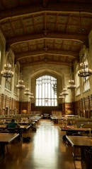 An ornate library hall with high, intricately designed ceilings and large arched windows allowing natural light to flood the space. Rows of wooden tables with lamps are arranged for study, and several individuals are seated, engrossed in work. The tall bookshelves line the sides, contributing to a scholarly atmosphere enhanced by the presence of large chandeliers providing a warm glow.