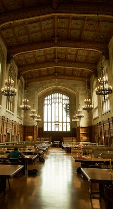 An ornate library hall with high, intricately designed ceilings and large arched windows allowing natural light to flood the space. Rows of wooden tables with lamps are arranged for study, and several individuals are seated, engrossed in work. The tall bookshelves line the sides, contributing to a scholarly atmosphere enhanced by the presence of large chandeliers providing a warm glow.