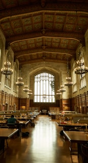 An ornate library hall with high, intricately designed ceilings and large arched windows allowing natural light to flood the space. Rows of wooden tables with lamps are arranged for study, and several individuals are seated, engrossed in work. The tall bookshelves line the sides, contributing to a scholarly atmosphere enhanced by the presence of large chandeliers providing a warm glow.