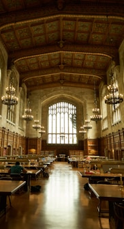 An ornate library hall with high, intricately designed ceilings and large arched windows allowing natural light to flood the space. Rows of wooden tables with lamps are arranged for study, and several individuals are seated, engrossed in work. The tall bookshelves line the sides, contributing to a scholarly atmosphere enhanced by the presence of large chandeliers providing a warm glow.