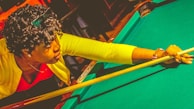 A person is playing pool, leaning over the table while aiming with a cue stick. The individual wears a bright yellow sweater and a red shirt, and appears focused on their shot. Their curly hair and a watch on the left wrist are visible.
