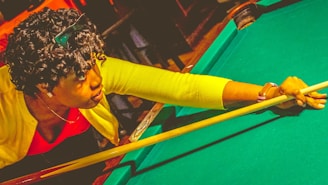 A person is playing pool, leaning over the table while aiming with a cue stick. The individual wears a bright yellow sweater and a red shirt, and appears focused on their shot. Their curly hair and a watch on the left wrist are visible.