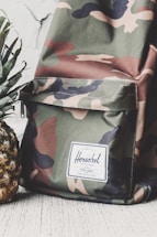 A camouflage-patterned backpack with a white label that reads 'Herschel' on the front pocket. The backpack is placed next to a fresh pineapple with green leaves. Both items are positioned against a light-colored, textured background.