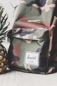 A camouflage-patterned backpack with a white label that reads 'Herschel' on the front pocket. The backpack is placed next to a fresh pineapple with green leaves. Both items are positioned against a light-colored, textured background.