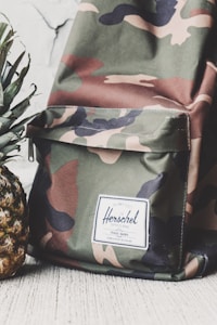 A camouflage-patterned backpack with a white label that reads 'Herschel' on the front pocket. The backpack is placed next to a fresh pineapple with green leaves. Both items are positioned against a light-colored, textured background.