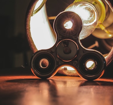 A metallic fidget spinner is positioned on a wooden surface, illuminated by a bright, glowing light bulb behind it. The light casts reflections and shadows, creating an industrial yet playful atmosphere.