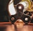 A metallic fidget spinner is positioned on a wooden surface, illuminated by a bright, glowing light bulb behind it. The light casts reflections and shadows, creating an industrial yet playful atmosphere.