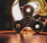 A metallic fidget spinner is positioned on a wooden surface, illuminated by a bright, glowing light bulb behind it. The light casts reflections and shadows, creating an industrial yet playful atmosphere.