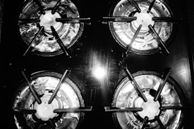 Four gas stove burners are arranged in a grid pattern. The burners are made of metal, showing signs of use with a slight shine from overhead lighting. The image is in black and white, emphasizing the textures and reflections on the metal surface.