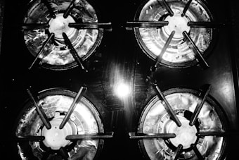 Four gas stove burners are arranged in a grid pattern. The burners are made of metal, showing signs of use with a slight shine from overhead lighting. The image is in black and white, emphasizing the textures and reflections on the metal surface.