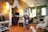 A cozy, stylishly decorated living room featuring brick and green walls with plenty of decorative elements. A young child wearing a navy blue sweatshirt and gray pants is standing in the center, raising an arm enthusiastically. The room includes furniture like a white chair, a small white table with a statue, and a desktop computer on a wooden desk. The ceiling has draped fabric and green plants, and there are multiple lamps providing warm lighting.
