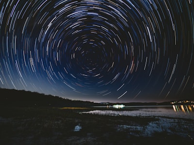 A stunning long-exposure photograph capturing a night sky filled with circular star trails, creating a hypnotic swirl pattern over a serene lake with a reflective surface. The horizon is lined with trees, and distant lights can be seen illuminating parts of the landscape, adding a sense of depth and contrast.