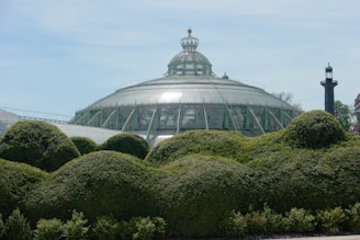 A large glass conservatory with a dome structure is surrounded by well-manicured green hedges. The conservatory is ornate with a detailed metal frame, and the sky is clear with soft blue and white hues. A tall black metal tower stands to the side, contrasting with the greenery.