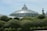 A large glass conservatory with a dome structure is surrounded by well-manicured green hedges. The conservatory is ornate with a detailed metal frame, and the sky is clear with soft blue and white hues. A tall black metal tower stands to the side, contrasting with the greenery.