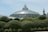 A large glass conservatory with a dome structure is surrounded by well-manicured green hedges. The conservatory is ornate with a detailed metal frame, and the sky is clear with soft blue and white hues. A tall black metal tower stands to the side, contrasting with the greenery.