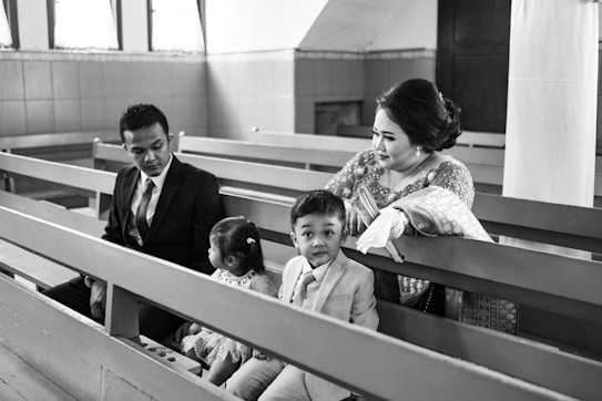 A family is seated on church pews. The scene includes a man in a suit, a woman in traditional attire, a young boy also dressed in a suit, and a girl in a dress. The woman and children seem engaged, with the man looking towards the girl.