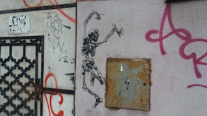 Graffiti art featuring a dancing skeleton on a textured wall surrounded by various tags and spray-painted text. The wall includes rusty metal elements and a patterned door grate on the left.