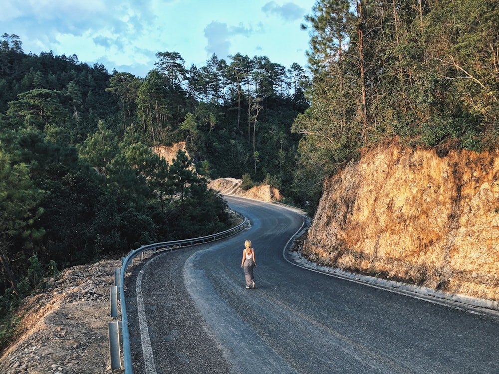 나무와 암석 사이에 회색 아스팔트 도로를 걷는 여자