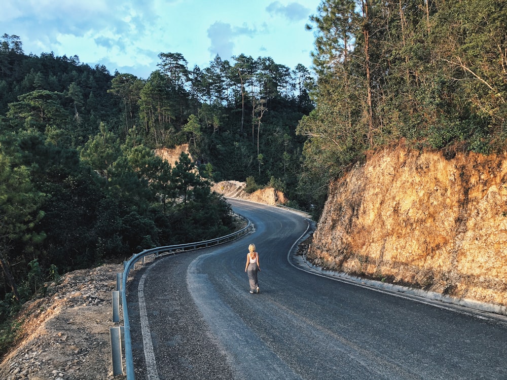 木々と岩層の間の灰色のアスファルト道路を歩く女性