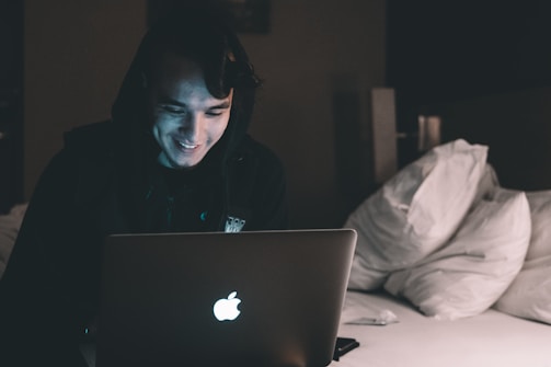 A person wearing a hoodie is sitting on a bed in a dimly lit room, focused on a laptop that is emitting a soft glow. Several pillows are stacked casually on the bed.