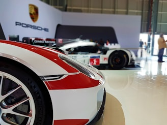 Two race cars, primarily white with red accents, are displayed in a well-lit indoor exhibition space. The foreground features part of a sports car, including the front wheel and headlight. The background shows the side profile of another car with a '1' racing number, and the Porsche logo is visible on the wall.