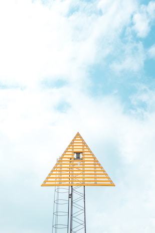 A tall metal structure supports a triangular, orange-striped sign against a bright, partly cloudy sky. The design is minimalist and modern, with the geometric lines of the sign standing out distinctly against the pale blue and white background.