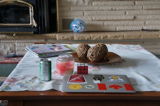 A wooden table with a white tablecloth is set with various items. There is a laptop adorned with stickers, a Rubik's cube, a lit pink candle, a green soda can, and a stack of colorful magazines. A decorative bowl containing three woven spheres sits in the center. In the background, there is a beige stone wall and a black fireplace.