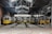 A series of yellow trams are parked inside a large, industrial tram depot with high ceilings and metal structures. The depot has visible rails leading to each tram, and there is natural light coming through the metal and glass roof. The trams are neatly aligned and display various signs on the front.