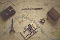 Various travel-related items are laid out on a map. The objects include a small model of the Eiffel Tower, a vintage camera, a modern instant camera, a notebook with a sketch of an architectural landmark, and a pencil. The map shows detailed streets and locations, likely from a city in Europe.
