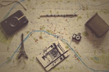 Various travel-related items are laid out on a map. The objects include a small model of the Eiffel Tower, a vintage camera, a modern instant camera, a notebook with a sketch of an architectural landmark, and a pencil. The map shows detailed streets and locations, likely from a city in Europe.