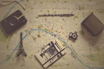 Various travel-related items are laid out on a map. The objects include a small model of the Eiffel Tower, a vintage camera, a modern instant camera, a notebook with a sketch of an architectural landmark, and a pencil. The map shows detailed streets and locations, likely from a city in Europe.