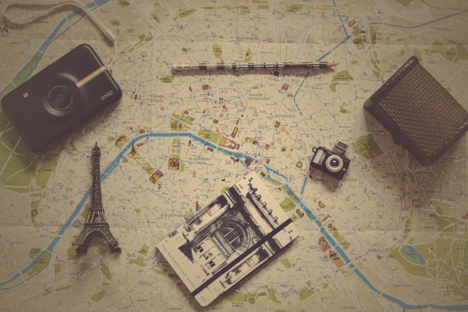 Various travel-related items are laid out on a map. The objects include a small model of the Eiffel Tower, a vintage camera, a modern instant camera, a notebook with a sketch of an architectural landmark, and a pencil. The map shows detailed streets and locations, likely from a city in Europe.