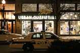 A storefront for Urban Outfitters is illuminated, showcasing various clothing items and accessories on display. In front of the store, a police car marked with 'Knoxville' is parked on the street. The scene is set at night, with a streetlamp casting light and autumn leaves on a tree visible to the side.