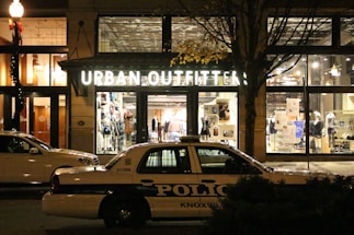 A storefront for Urban Outfitters is illuminated, showcasing various clothing items and accessories on display. In front of the store, a police car marked with 'Knoxville' is parked on the street. The scene is set at night, with a streetlamp casting light and autumn leaves on a tree visible to the side.