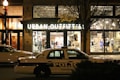A storefront for Urban Outfitters is illuminated, showcasing various clothing items and accessories on display. In front of the store, a police car marked with 'Knoxville' is parked on the street. The scene is set at night, with a streetlamp casting light and autumn leaves on a tree visible to the side.