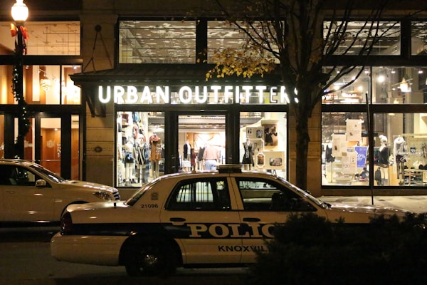 A storefront for Urban Outfitters is illuminated, showcasing various clothing items and accessories on display. In front of the store, a police car marked with 'Knoxville' is parked on the street. The scene is set at night, with a streetlamp casting light and autumn leaves on a tree visible to the side.