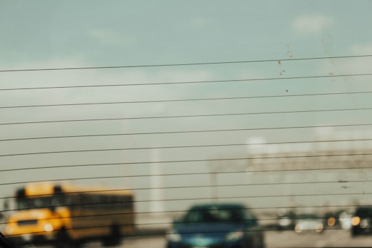 A blurred view through the back window of a vehicle, featuring horizontal defrost lines. In the background, there is a yellow school bus and several cars driving on a highway, all slightly out of focus. The sky is partially cloudy with a faint blue tint.