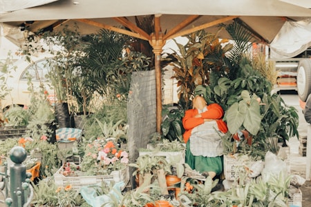 A vendor is relaxing under a large umbrella surrounded by a variety of plants and flowers. The scene is bustling with greenery, creating a dense, lush atmosphere. The vendor appears to be resting, wearing a bright red jacket and hat. The market stall is filled with different types of potted plants, some of which have vibrant flowers.