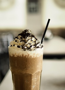 A tall, frothy chocolate drink topped with whipped cream and sprinkled with crushed cookies. A black straw is placed in the drink, which is served in a clear glass with condensation on the outside.