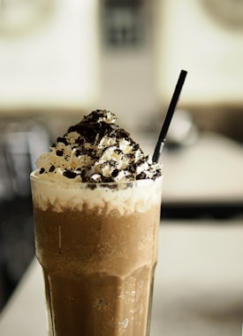 A tall, frothy chocolate drink topped with whipped cream and sprinkled with crushed cookies. A black straw is placed in the drink, which is served in a clear glass with condensation on the outside.
