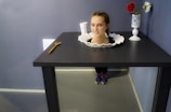 A playful optical illusion where a woman's head appears to be on a plate set in the middle of a black table, with her legs hidden behind the table. Nearby, there is a vase with two flowers and a white cup, as well as a small wooden object resembling a wrench.