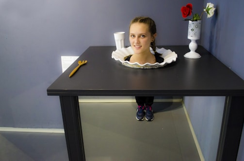 A playful optical illusion where a woman's head appears to be on a plate set in the middle of a black table, with her legs hidden behind the table. Nearby, there is a vase with two flowers and a white cup, as well as a small wooden object resembling a wrench.