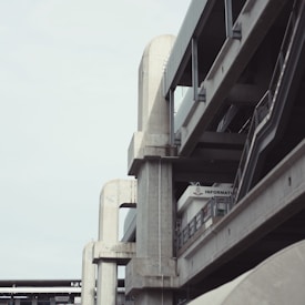 A modern concrete structure with multiple levels, featuring prominent vertical and horizontal lines. The structure has an industrial design, with a sky backdrop and an information sign visible. The architecture displays exposed beams and support columns that are robust and symmetrical.