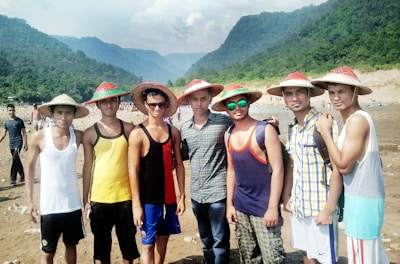 A group of young men stand together, wearing straw hats. They are dressed in casual summer attire, including tank tops and sunglasses. The background features a scenic view of mountains and lush greenery under a cloudy sky, with a rocky riverbank visible.