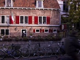 A charming, historic brick building with red shutters and a red-tiled roof sits in a European street setting. Bicycles are parked along the canal in front, and small potted plants line the stone edge. An Italian flag hangs on the left side of the building, indicating the presence of an Italian restaurant.