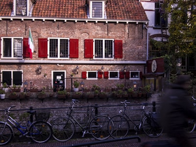 A charming, historic brick building with red shutters and a red-tiled roof sits in a European street setting. Bicycles are parked along the canal in front, and small potted plants line the stone edge. An Italian flag hangs on the left side of the building, indicating the presence of an Italian restaurant.
