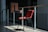 A red plastic chair with a wooden and metal desk attachment is positioned in a sunlit corridor. The scene shows a contrast between light and shadows, with the chair catching the sunlight. The surroundings include a concrete floor and metal railings, contributing to an industrial atmosphere.