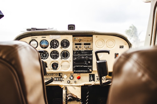 A detailed view of an airplane cockpit featuring a variety of dials, gauges, and controls. The image captures the instrument panel with indicators for altitude, speed, and other flight metrics. The cockpit is framed by the two pilot seats upholstered in brown leather.