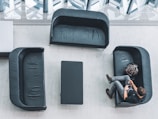 An overhead view of a modern waiting area with three black leather sofas arranged around a rectangular black table on a light gray floor. A person is seated on the right sofa, looking at a phone, with a backpack placed beside them.